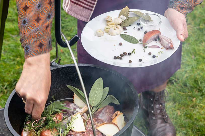 Pan met vlees boven een houtvuurtje, iemand voegt verse kruiden toe.